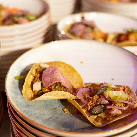 Two tacos on a white plate with a blue and brown rim, filled with slices of meat, chopped onions, pickled red onions, green peppers, and a crumbly topping. A stack of similar plates is visible in the background.