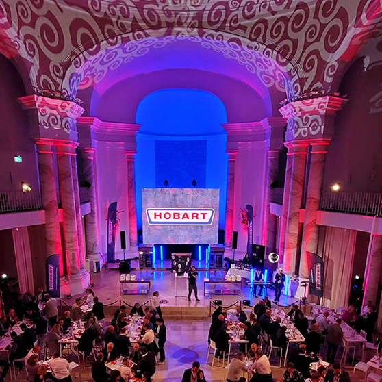 Large event hall with high ceilings and elegant architecture, illuminated in shades of purple, blue, and red. A central stage features a large screen displaying the word “HOBART”. Numerous guests are seated at round tables, suggesting a festive event or conference setting.