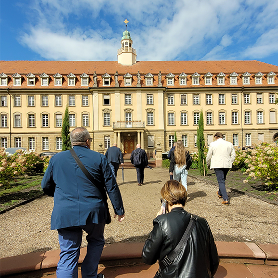 A group of people walks toward a large historic building with a red-tiled roof and a central tower featuring a clock and a cross. The path is lined with well-maintained gardens filled with flowers and shrubs. The scene takes place during daylight under a partly cloudy sky.