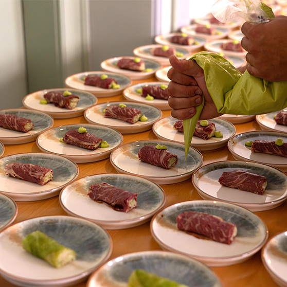 A person uses a piping bag to apply green garnish or sauce onto multiple plates, each containing a rolled piece of meat with filling. The plates are arranged in rows on a table, suggesting preparation for a large culinary event.