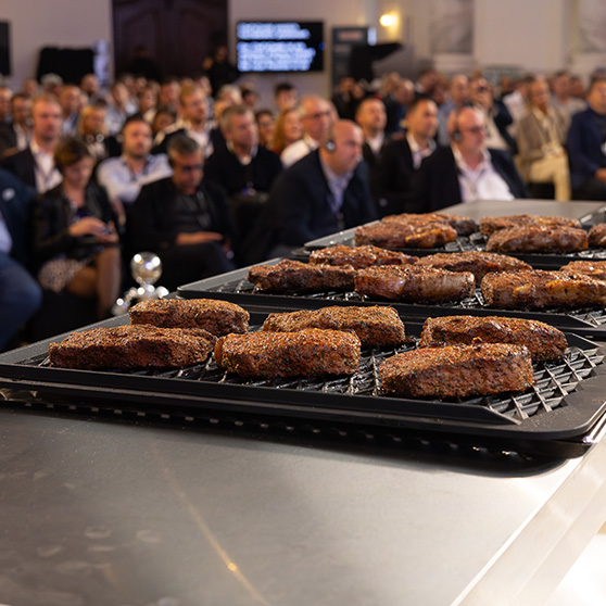 Grilled steaks on a tray during a live cooking presentation in front of a large audience