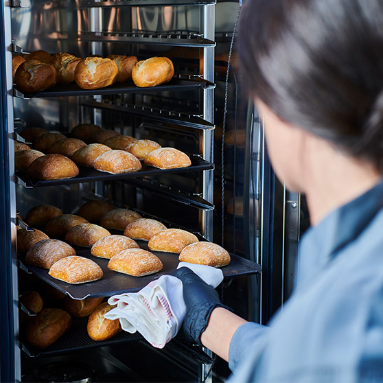 A person removes freshly baked bread rolls from Hobart's CHEF'S COMBI using gloves and a cloth – multiple trays of golden-brown rolls in a professional oven.