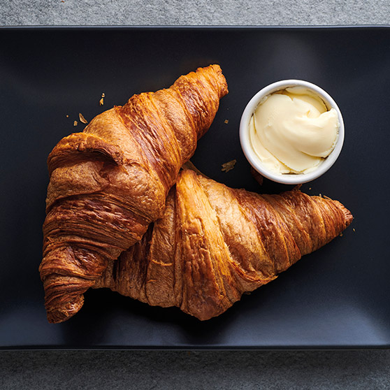 Two golden-brown, freshly baked croissants on a black plate with a bowl of butter – prepared using HOBART's CHEF'S COMBI for perfect texture and browning.