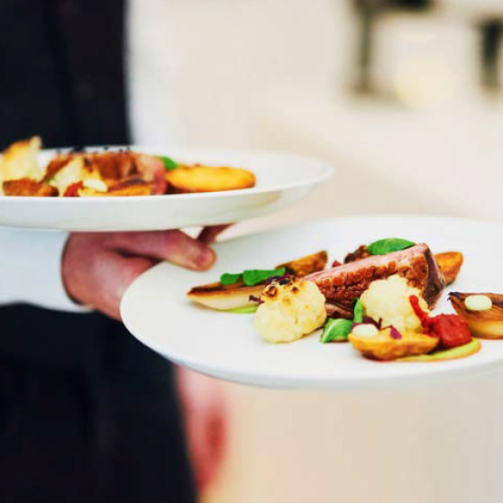 A person holds two elegantly plated dishes with meat, roasted vegetables, and refined garnishes – prepared using HOBART's CHEF'S COMBI for top-level professional kitchen standards.