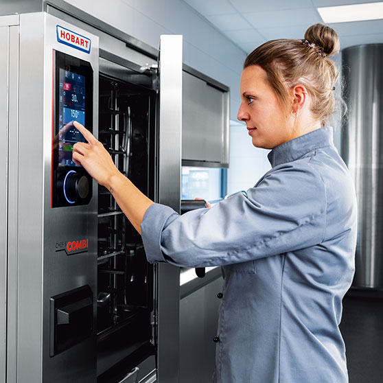 A person operates the touchscreen display of a CHEF'S COMBI unit – showing time and temperature settings like “30” and “150°”, enabling precise control in professional kitchen environments.