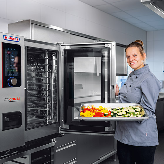 Professional HOBART combi steamer in a modern stainless steel commercial kitchen, door open, tray with fresh vegetables including peppers and zucchini being loaded for efficient cooking in foodservice operations