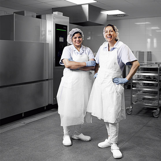 Two staff members in hygiene clothing standing in the dishwashing area of Heinsberg hospital in front of an industrial dishwasher with stainless steel trolleys and modern hygiene technology for efficient cleaning