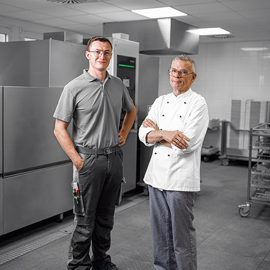 A technician and a chef standing in the dishwashing area of Heinsberg hospital in front of an industrial tray dishwasher with stainless steel trolleys and modern hygiene technology for efficient cleaning