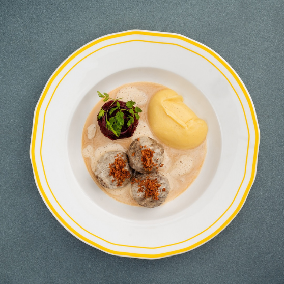 White plate with yellow rim on a gray table, serving Königsberger meatballs with creamy white sauce and side dish, traditionally plated.