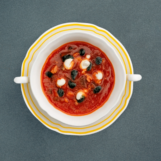 White soup bowl with Soljanka, a hearty Eastern European tomato-based soup topped with sour cream and black olives, served on a striped yellow plate