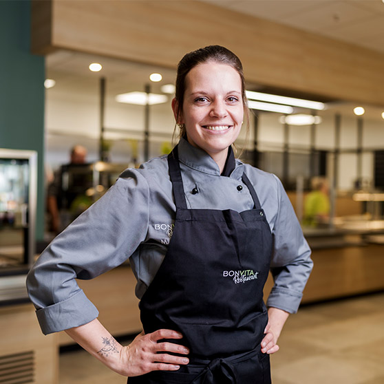 Staff member wearing a chef jacket and apron standing in a modern company kitchen. Serving counters and stainless steel furniture are visible in the background.