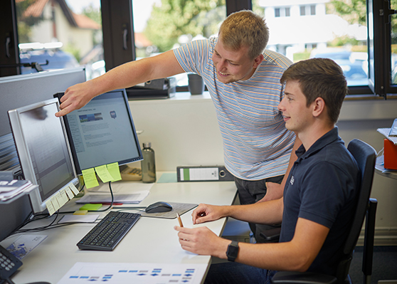 Apprenticeship setting in a modern office: trainee and instructor working together at a computer, discussing content on the screen and using notes for training. Desk includes keyboard, mouse, sticky notes, and writing tools.