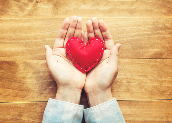 Two open hands holding a red, hand-stitched felt heart on a wooden surface. Represents love, care, craftsmanship, and appreciation.